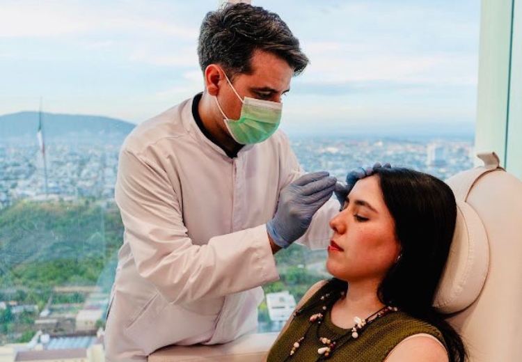 A woman in medical clothing applies a facial mask to a man's face in an indoor healthcare setting, surrounded by medical equipment and supplies.