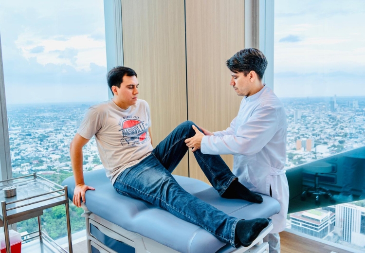A man wearing a black shirt and black pants is lying on a blue medical bed inside a healthcare room. The setting includes various pieces of medical equipment and clean furnishings.