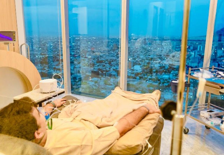 A group of people sitting in chairs in a living room, with a couch and artwork on the walls. The individuals are dressed in various clothing styles, and there is furniture scattered around the floor.