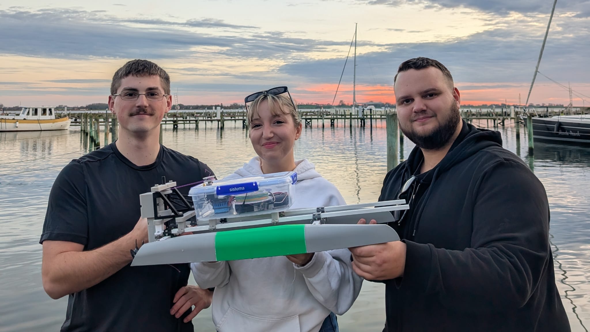 Three members of the Krakens team stand on a marina dock at sunset, smiling and holding a small prototype trimaran boat with a green hull section and a transparent plastic electronics box, with calm water, moored boats, and a pastel sky in the background.