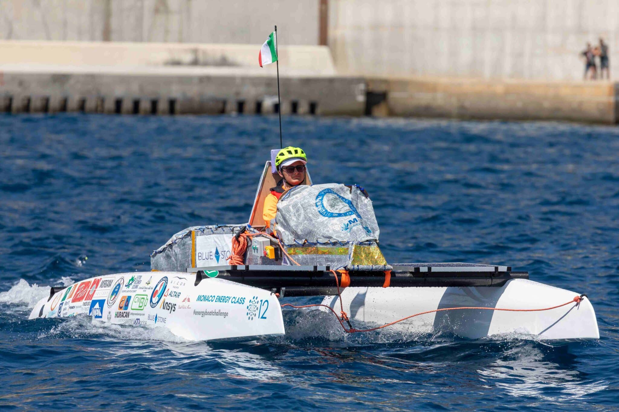 A single-seat, sponsor-covered experimental boat from the Monaco Energy Boat Challenge moves across deep blue water, piloted by a person in an orange life vest and bright yellow helmet, with an Italian flag flying from a thin mast and a concrete harbor wall and a few distant spectators in the background.