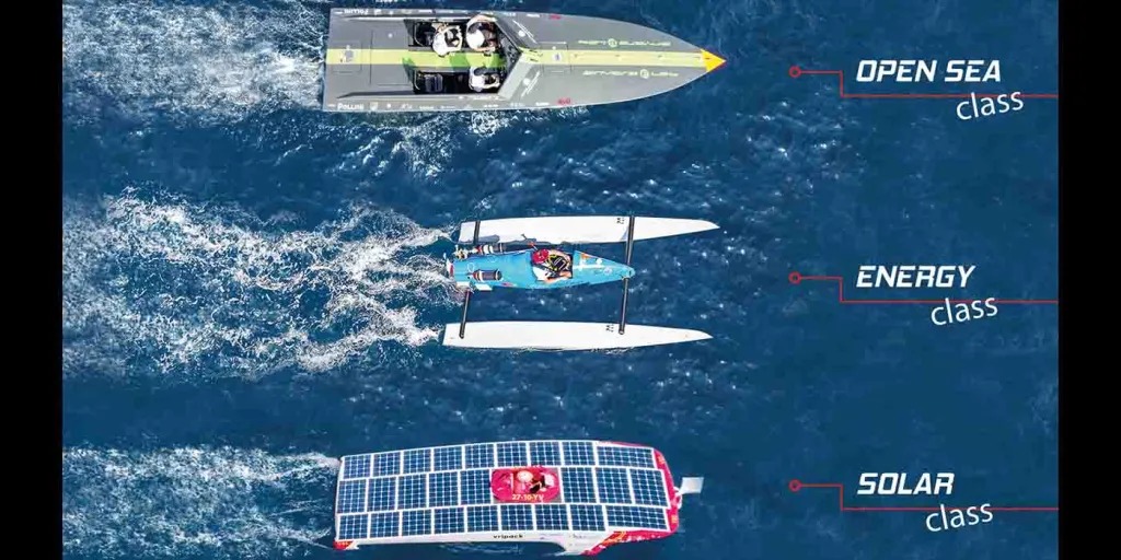 A single-seat, sponsor-covered experimental boat from the Monaco Energy Boat Challenge moves across deep blue water, piloted by a person in an orange life vest and bright yellow helmet, with an Italian flag flying from a thin mast and a concrete harbor wall and a few distant spectators in the background.