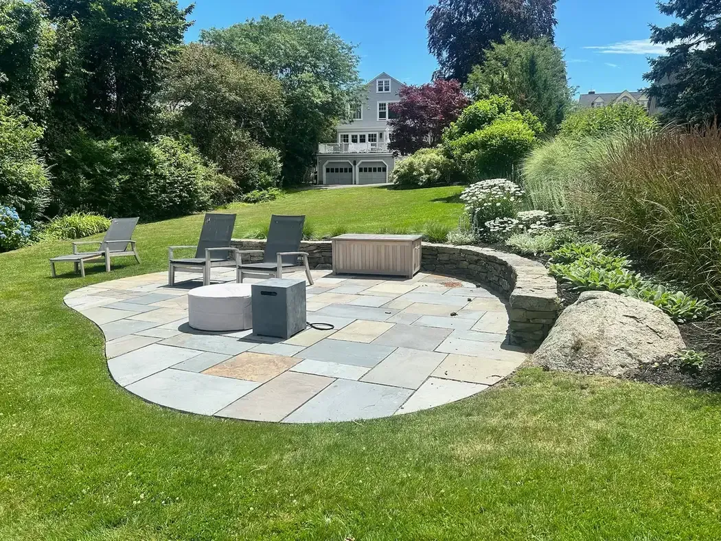 Custom bluestone patio with curved natural stone sitting wall and professionally landscaped backyard in Marblehead, MA by Nash & Brown Masonry.