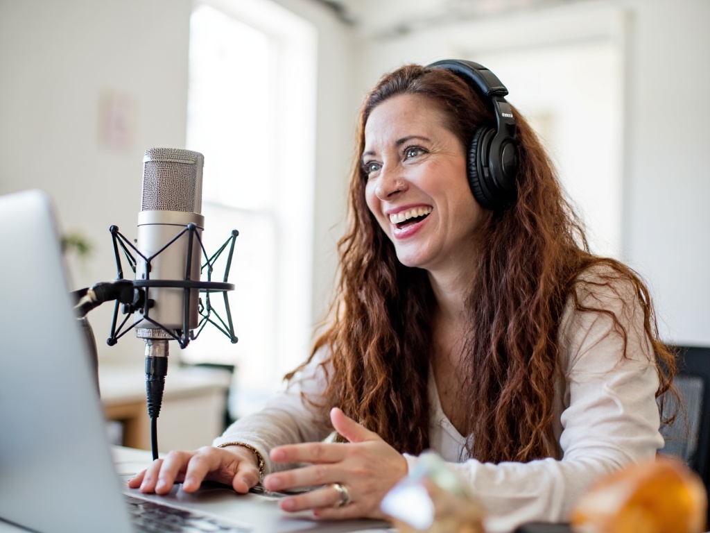 Professional woman in a modern studio, wearing headphones and speaking confidently into a microphone, with soft lighting and a blurred background of audio equipment, conveying expertise and luxury podcasting environment.