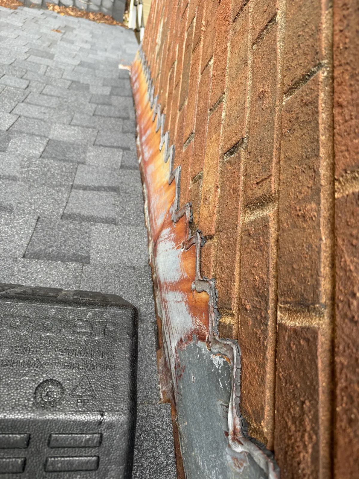 Close-up of metal step flashing along the side of a brick chimney meeting a shingled roof.