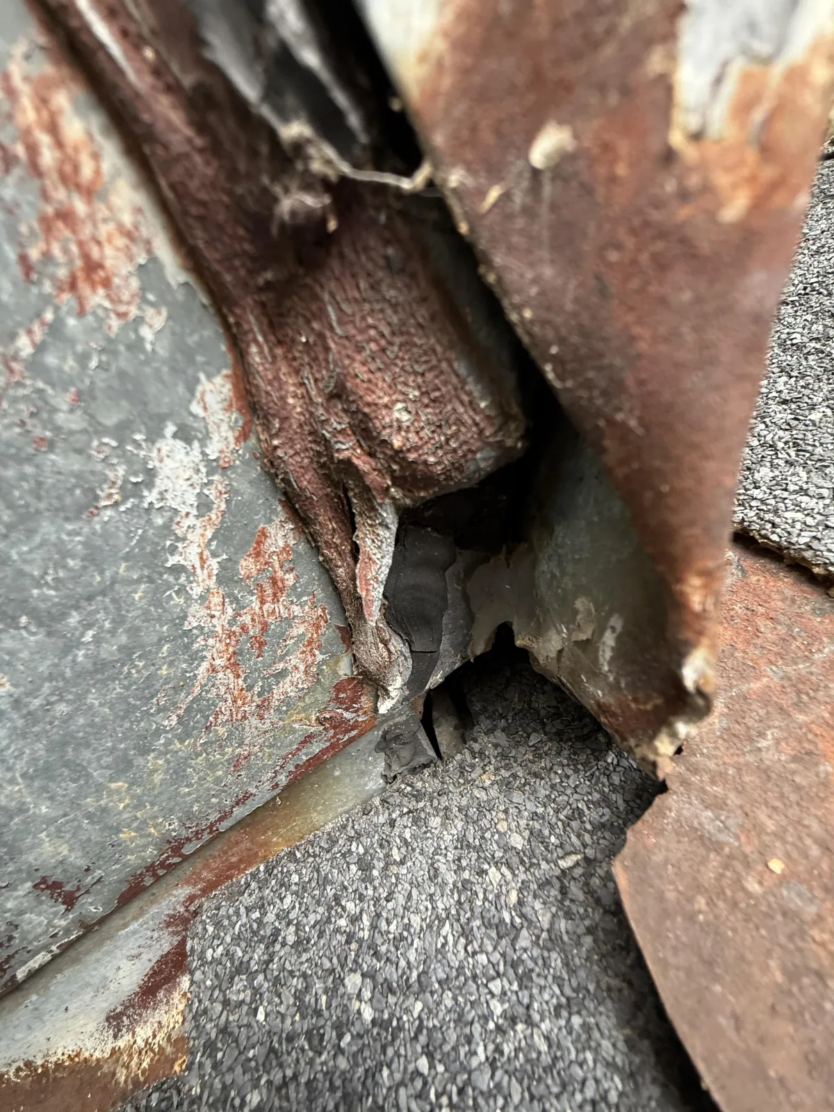 Close-up of severely rusted metal flashing and rotting material at a roof and chimney junction.