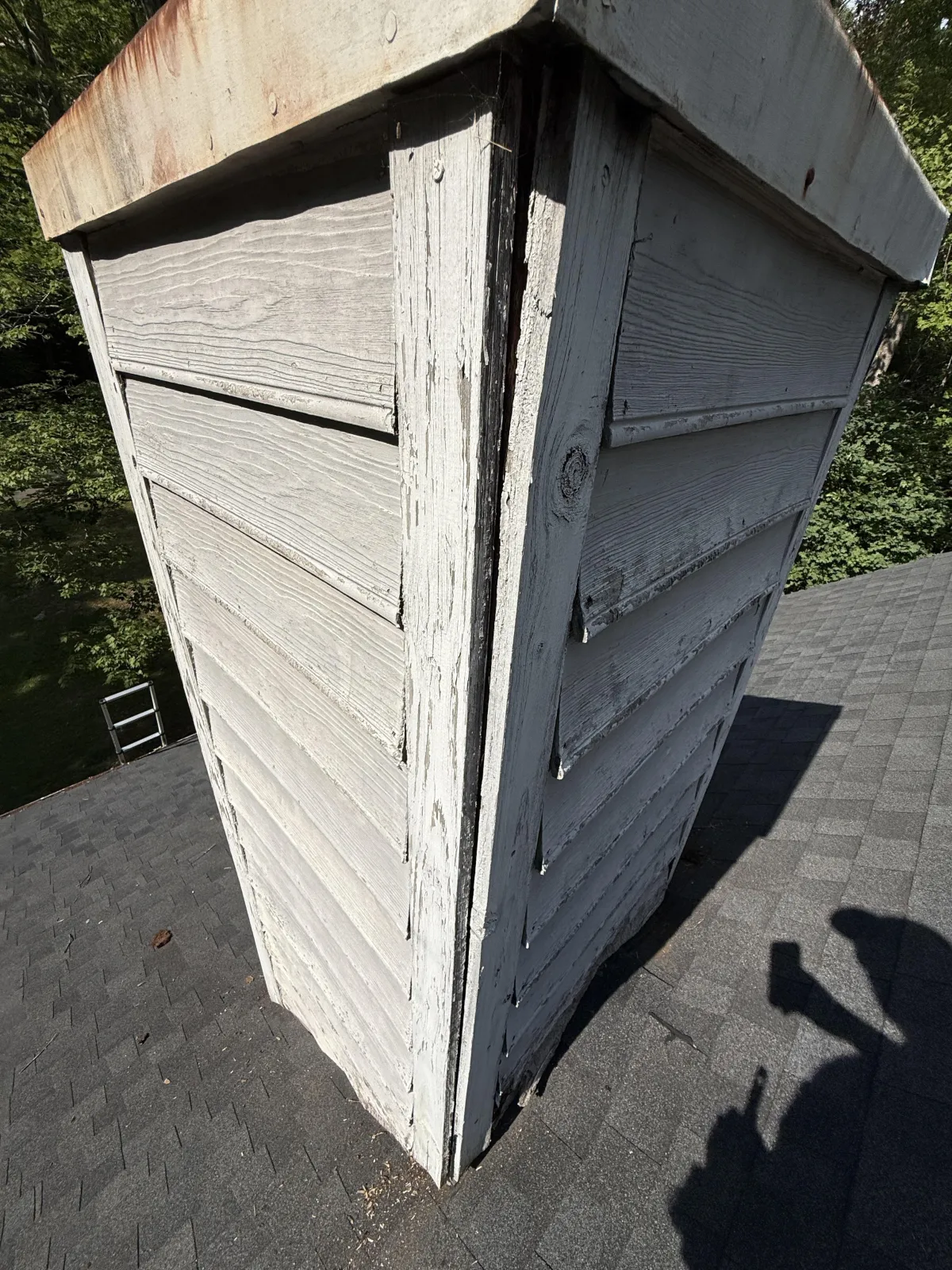 Close-up of a weathered chimney chase cover and peeling white horizontal siding panels on a shingle roof.