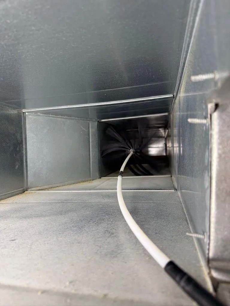 Interior view of a rectangular metal HVAC duct being cleaned with a rotary brush system.