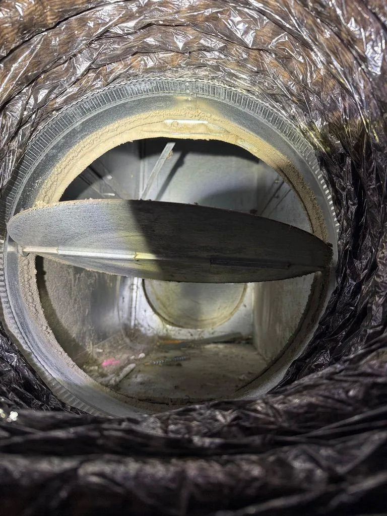 Interior view looking through a flexible HVAC duct at a metal damper valve with dust accumulation