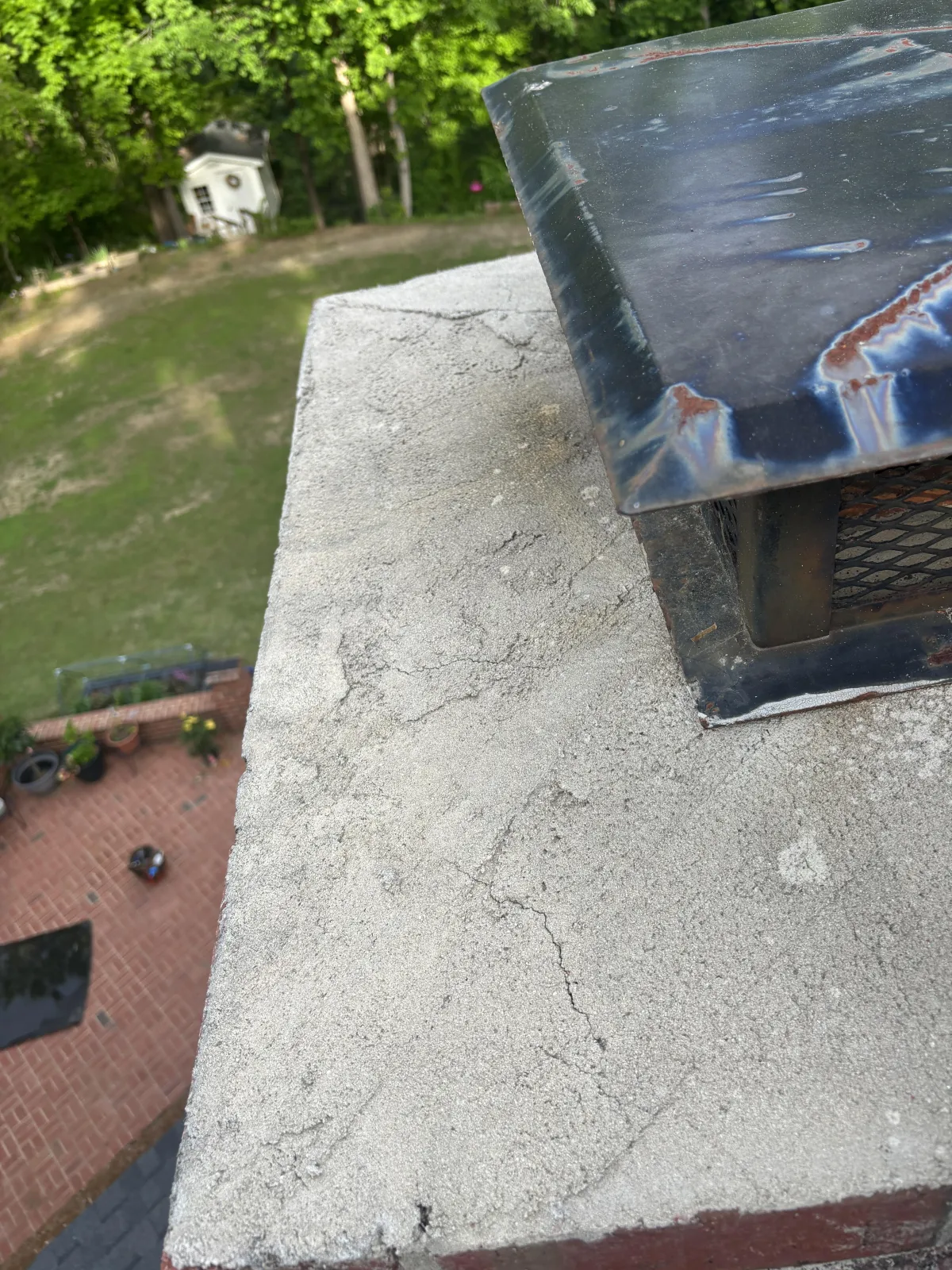 High-angle view of a damaged, cracked concrete chimney crown next to a metal flue cap