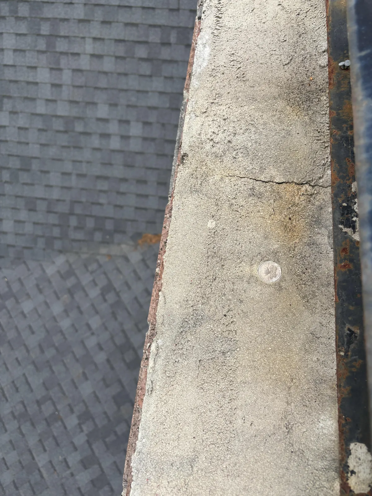 Close-up inspection view of a weathered concrete parapet wall with hairline cracks and rust next to a shingled roof