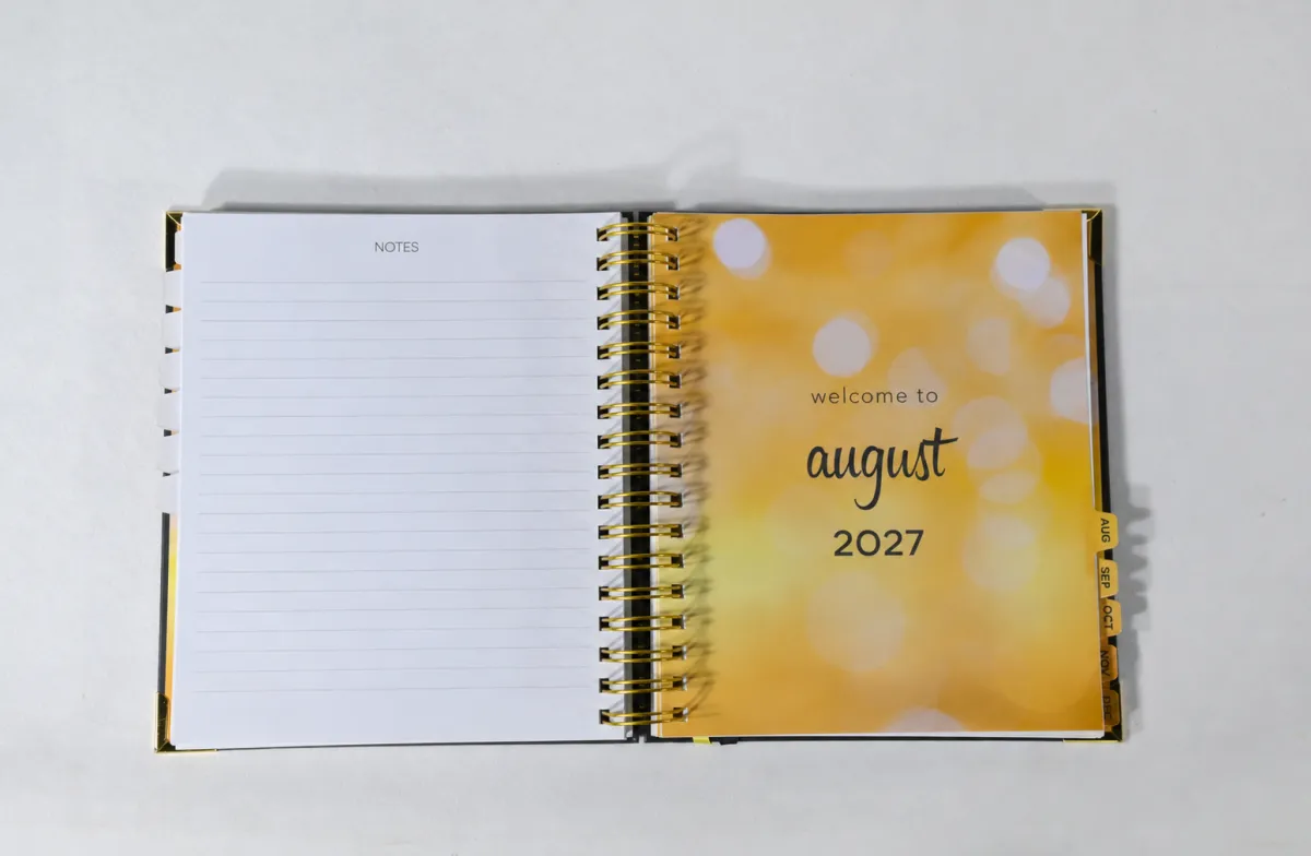 Hands writing with a silver pen in an open planner on a sunlit wooden desk