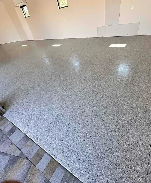 A newly installed light gray epoxy garage floor with speckled finish, reflecting light, next to dark patio pavers.