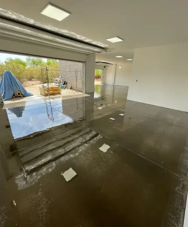 A newly constructed garage with a glossy, reflective dark floor, square ceiling lights, and an open garage door revealing an outdoor desert landscape.