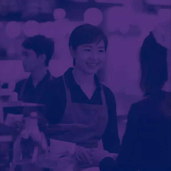 A barista wearing an apron smiling while interacting with a customer across the counter, shown in a purple-tinted photographic style.