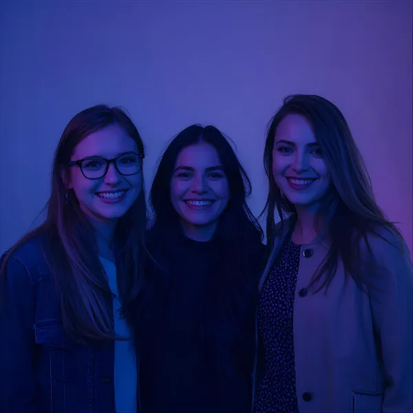 Three young women standing close together and smiling, illuminated by soft blue and purple lighting against a plain background.