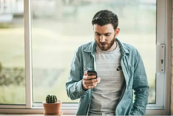 Man in casual attire responding to customer reviews for his local business on Myracai.com on his smartphone, standing by a window with a cactus plant nearby.