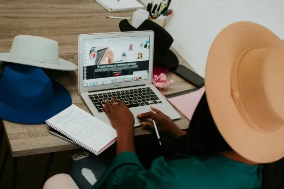 Person working on a laptop with stylish hats on the desk, exploring Myracai.com for creating a website for her small business..