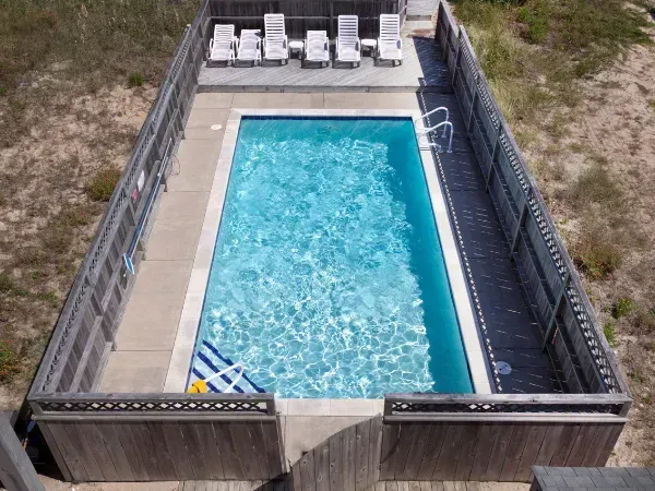 Rectangular swimming pool surrounded by a tall wooden privacy fence with lounge chairs in the background.