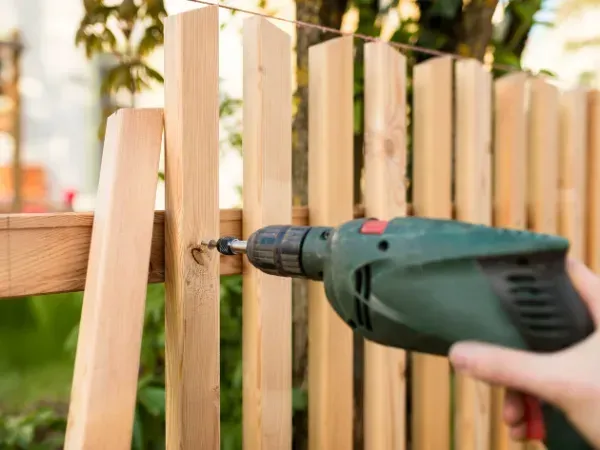 Close-up of a person using a power drill to attach wooden planks during fence installation.