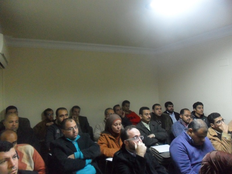 A group of people sitting at desks in a classroom or seminar, viewed from the back.