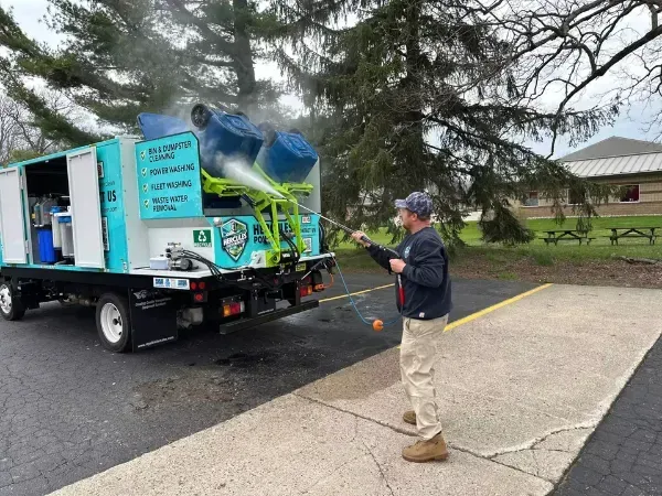 Operator pressure washing a blue bin on a Hercules cleaning truck in a tree-lined lot