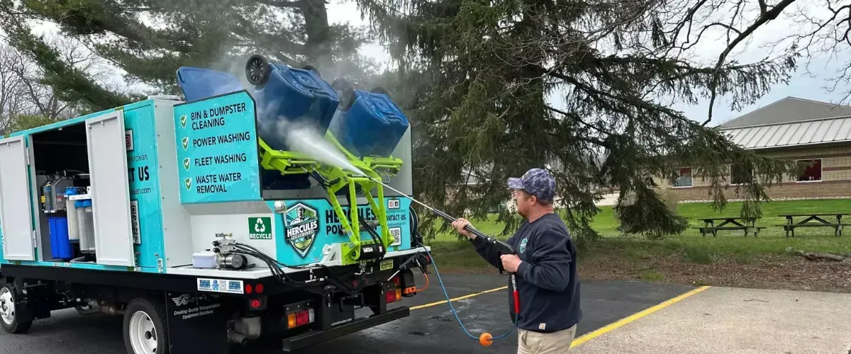 Worker pressure washing two blue dumpster bins lifted on a cleaning truck