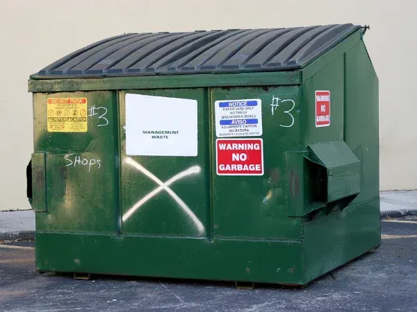Green commercial dumpster with warning signs and graffiti markings on the front.
