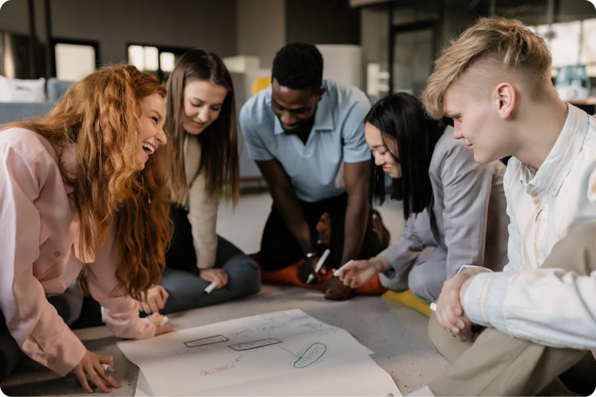 Diverse team kneeling on the floor, smiling and sketching a flowchart on large paper during a strategy session in a modern office.