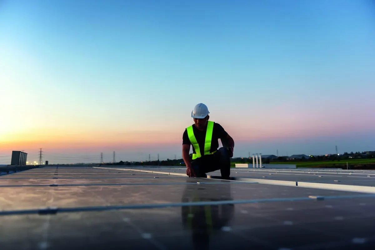 a person in a reflective vest and white hard hat kneeling on a shiny roof