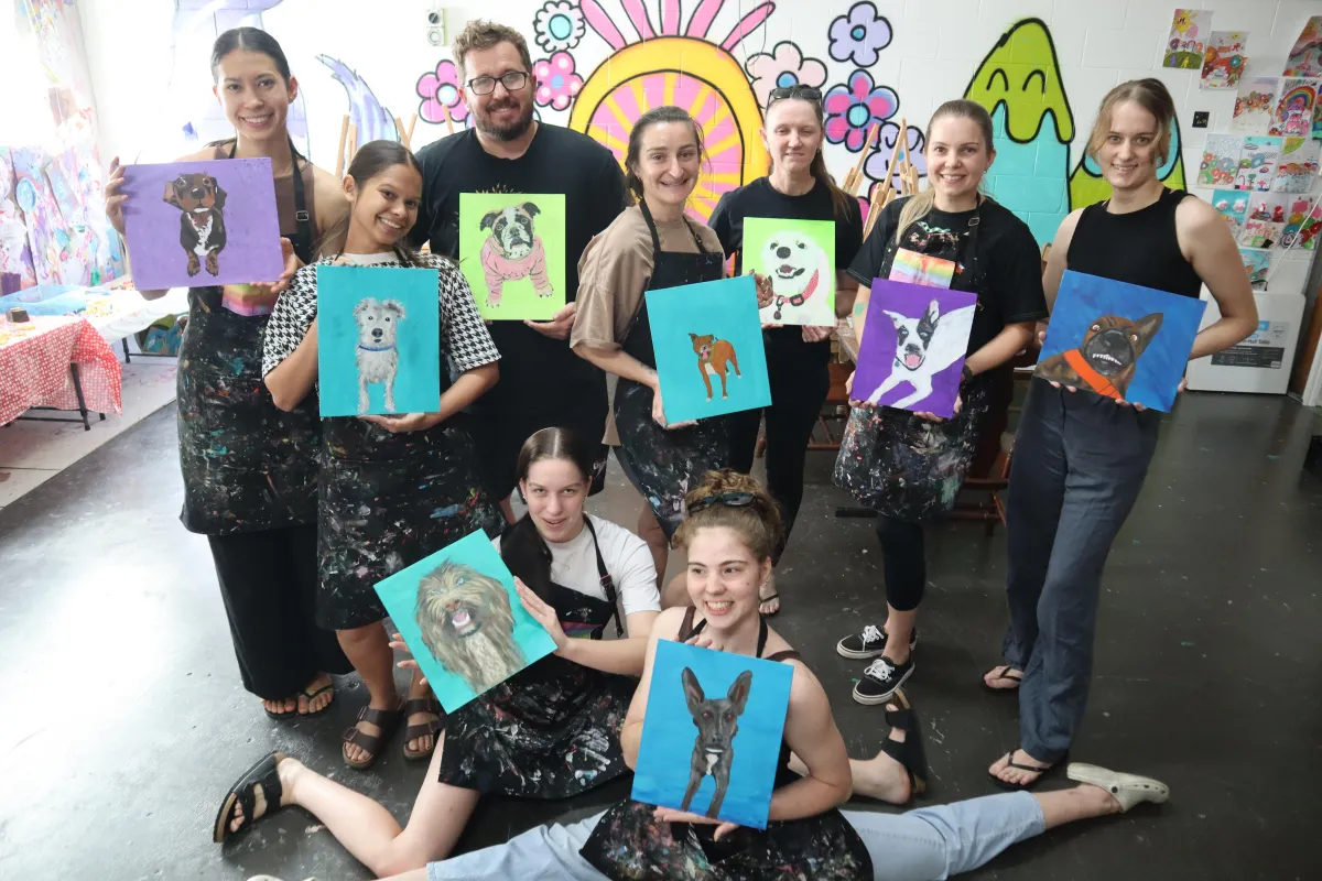 Photo of a group of colleagues in business casual attire, smiling and holding up their finished paintings in a pastel-decorated studio, with paint palettes and easels visible.