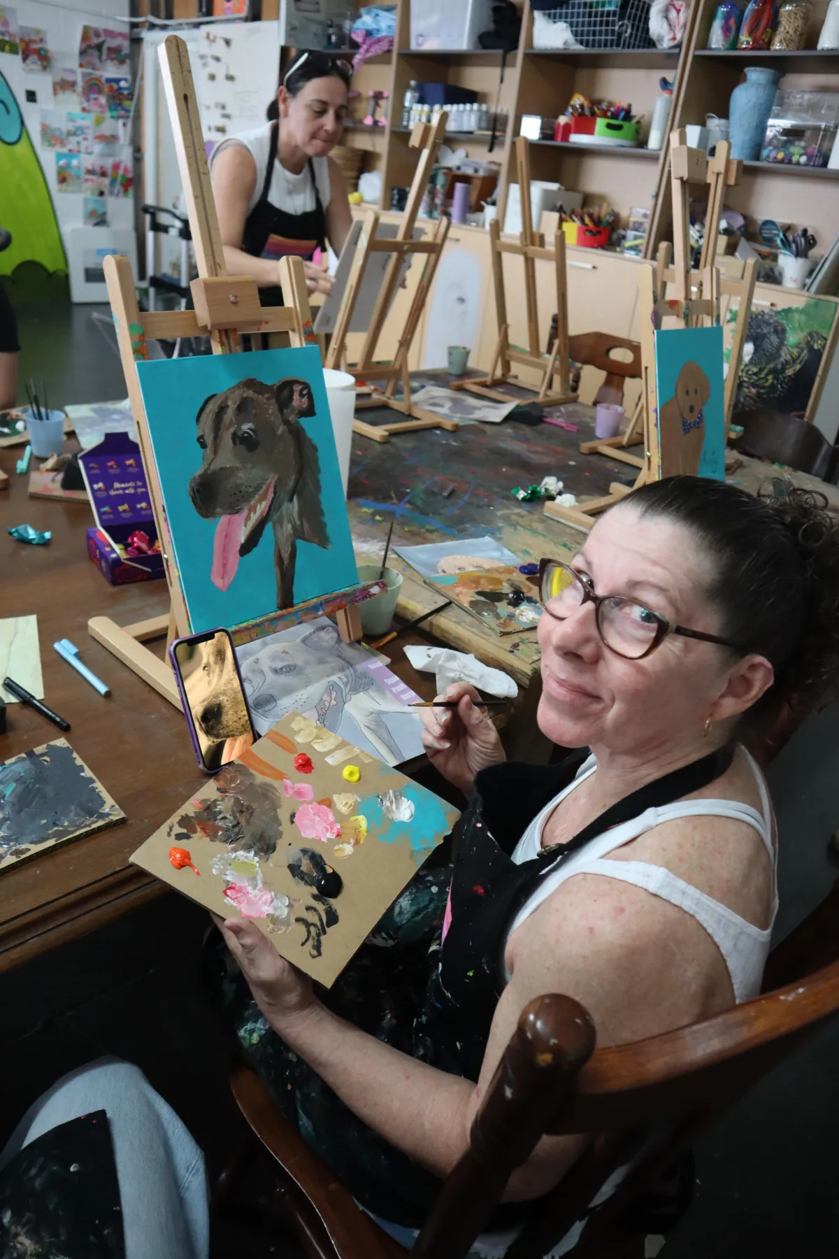 Photo of a professional artist demonstrating a painting technique to a group of attentive employees, with colourful canvases and wine glasses on the tables, in a bright, airy space.