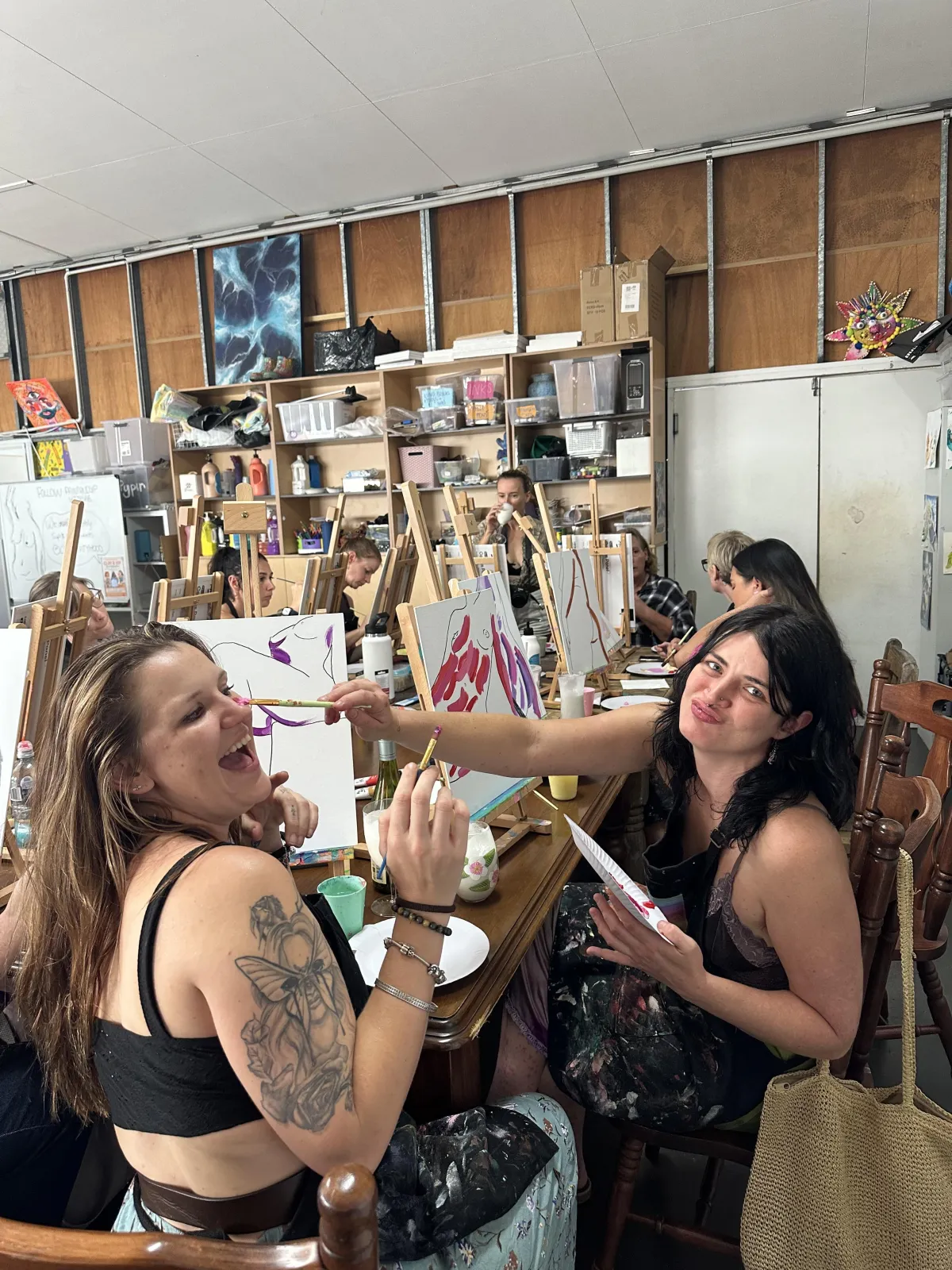 Photo of a diverse group of coworkers clinking wine glasses and laughing, surrounded by art supplies and half-finished paintings, with pastel bunting in the background.