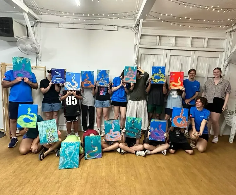 Wide workshop scene: hands-on mixed-media table with bright craft materials and instructor guiding children, lively composition, no faces close-up.