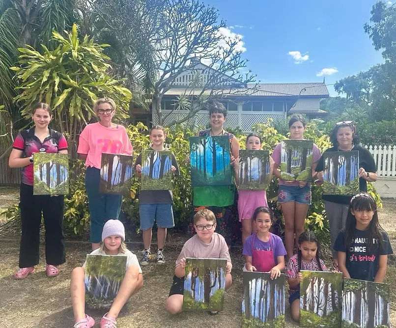 Wide workshop scene: hands-on mixed-media table with bright craft materials and instructor guiding children, lively composition, no faces close-up.