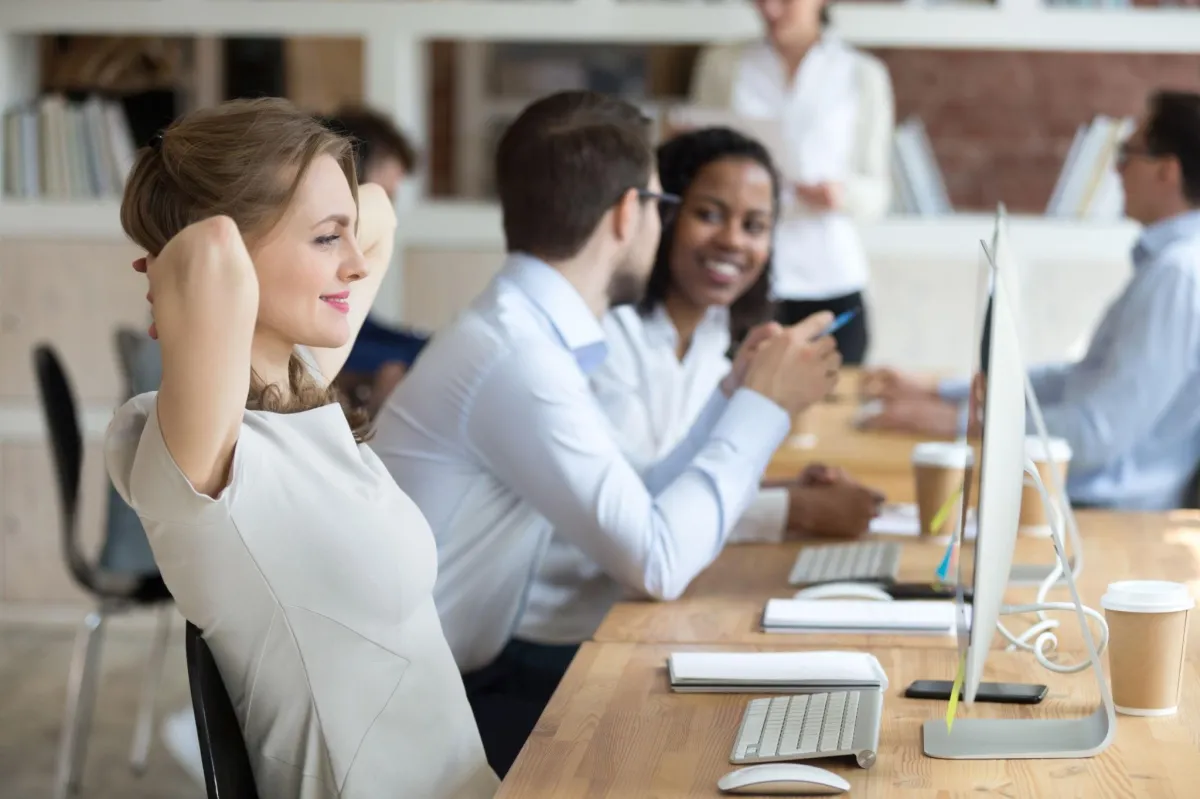 a happy and relaxed lookin woman sittin gat a computer desk with coworkers talking in the backround