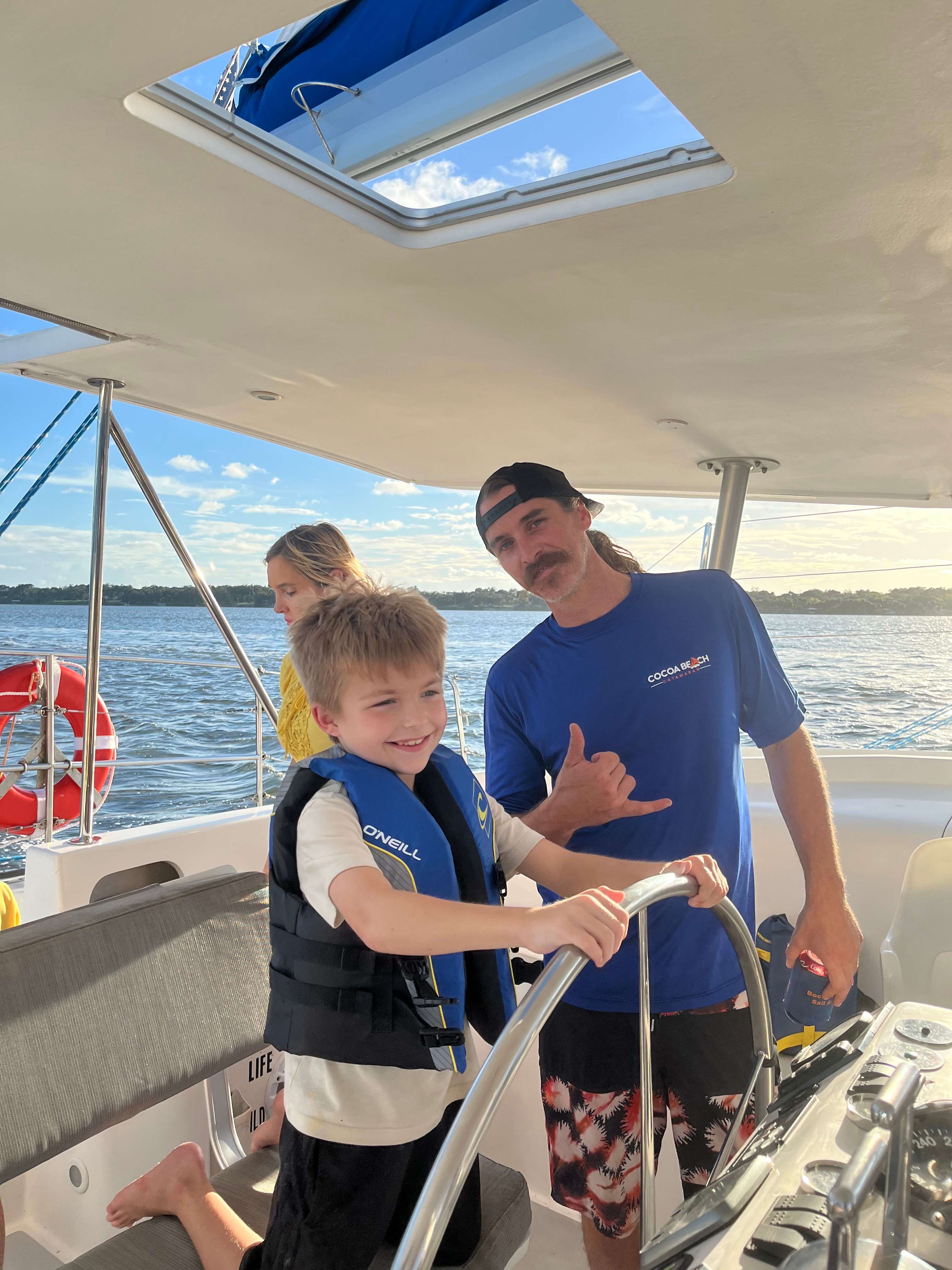 Child steering a catamaran with a smiling captain, both enjoying a dolphin adventure sail experience in Cocoa Beach, showcasing personalized sailing and wildlife encounters.