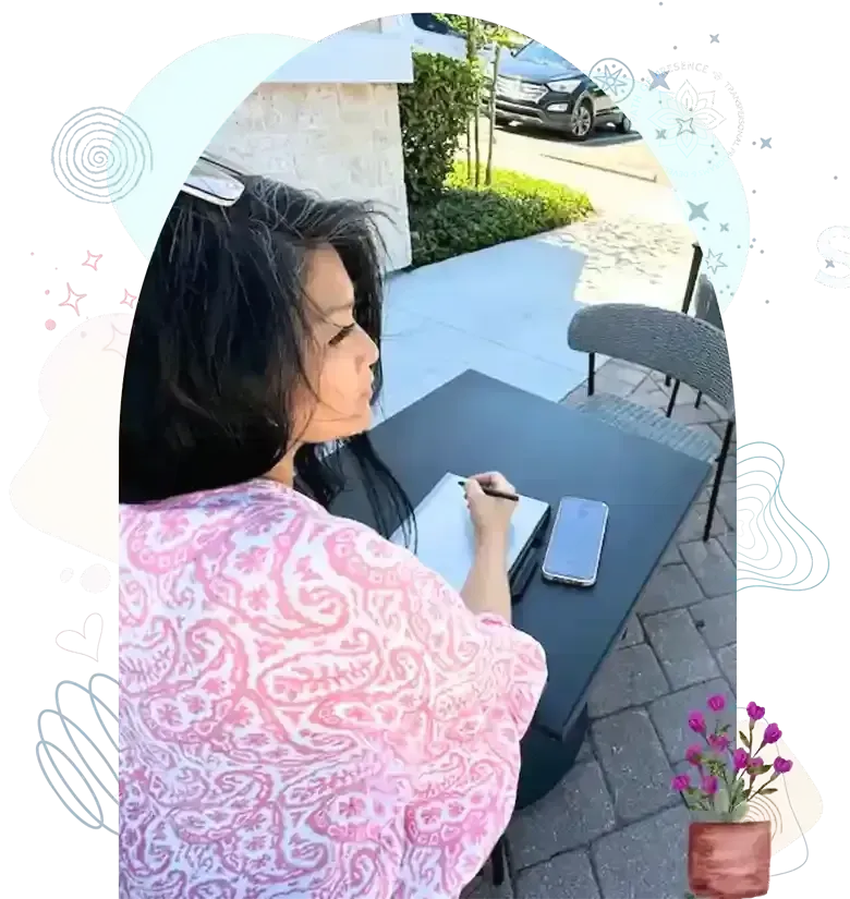 Person in a pink patterned top writing in a notebook at an outdoor table with a smartphone.