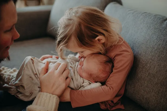 A Toddler holds their newborn sibling while mom watches