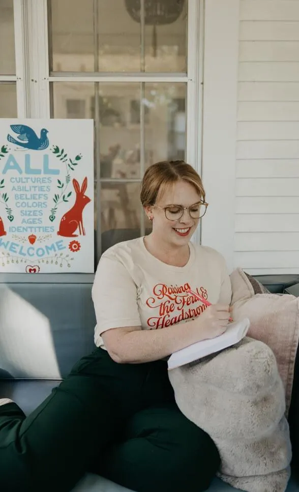 A smiling person with glasses sits on a couch, writing in a notebook. Behind them is a colorful sign with illustrations of animals and text welcoming all cultures, abilities, and identities in this body positive, social justice space.