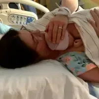 A new mom lies in a hospital bed, holding her newborn baby wrapped in a blanket to her chest as they both rest peacefully after birth.