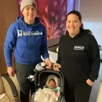 Two smiling queer women stand beside a newborn baby in a car seat. They are about to leave the hospital. One wears a blue University of Baltimore hoodie and gray beanie, the other a black Maui hoodie. Captured indoors in a cozy room, this sweet moment reflects the joy of new parenthood.