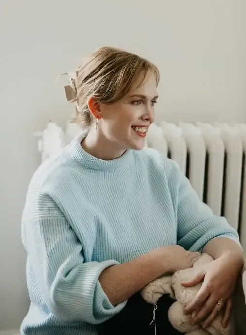 A woman with light hair tied back in a bow sits smiling, wearing a light blue sweater and holding a ball of beige yarn—radiating calm, she is ready for birth prep or prenatal support near a white radiator and neutral background.