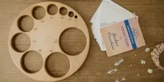 A round wooden tray demonstrate with numbered circular slots sits on a wooden cervical dilation for the purpose of birth education. It's next to a stack of cards and an Evidence Based Birth Pocket Book, alongside a crystal bead ornament. 