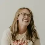 A young woman with light brown hair and glasses is smiling and laughing. She is wearing a light-colored shirt with red text, sitting against a plain background—Jillian Carpenter, an experienced birth doula offering compassionate birth support.