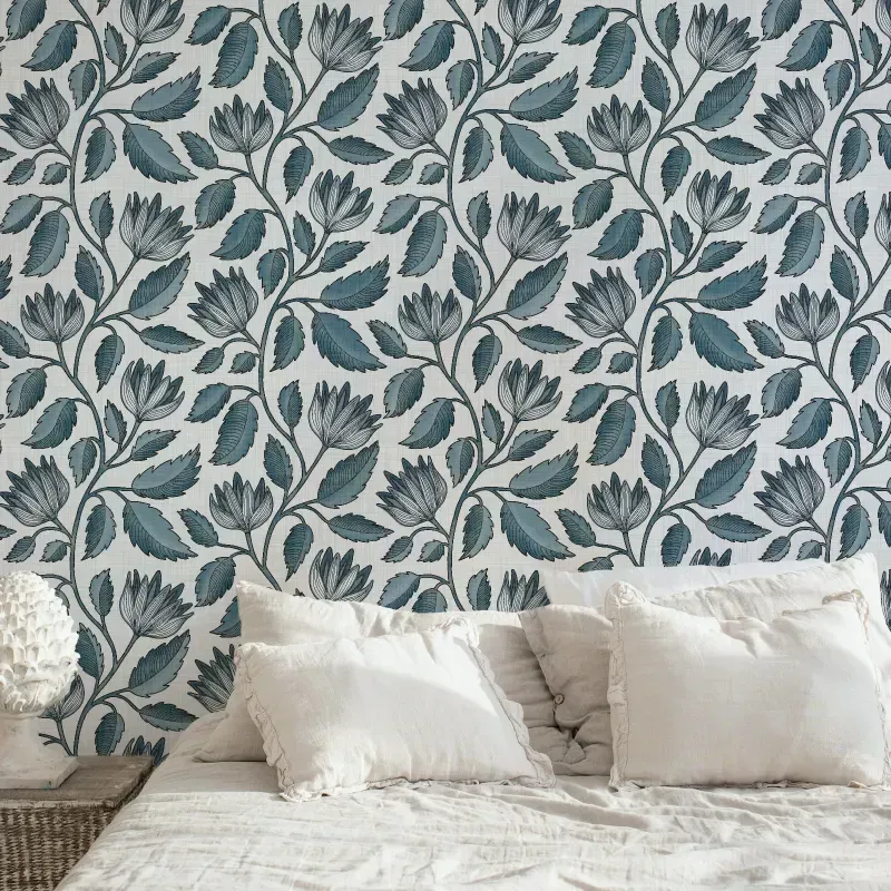 Blue hand drawn botanical floral wallpaper with leafy vines and outlined flowers, styled behind a bed with cream pillows in a bedroom.