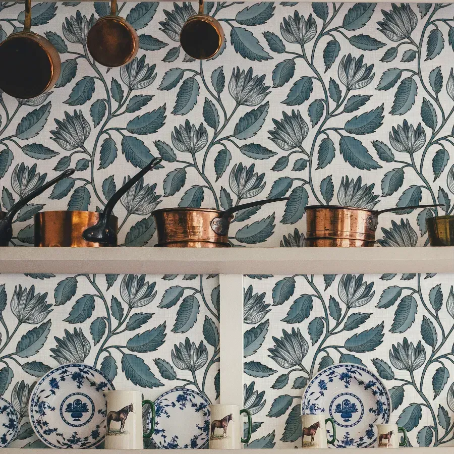Hand drawn blue botanical floral wallpaper with leafy vines and outlined flowers, styled behind kitchen shelves with copper pots and plates.