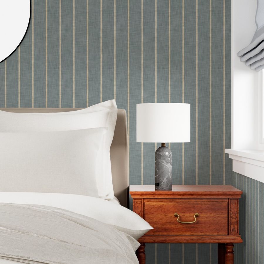 Slate blue-grey Roselk Design wallpaper with narrow beige stripes behind white bedding and a wooden bedside table in a bright bedroom.