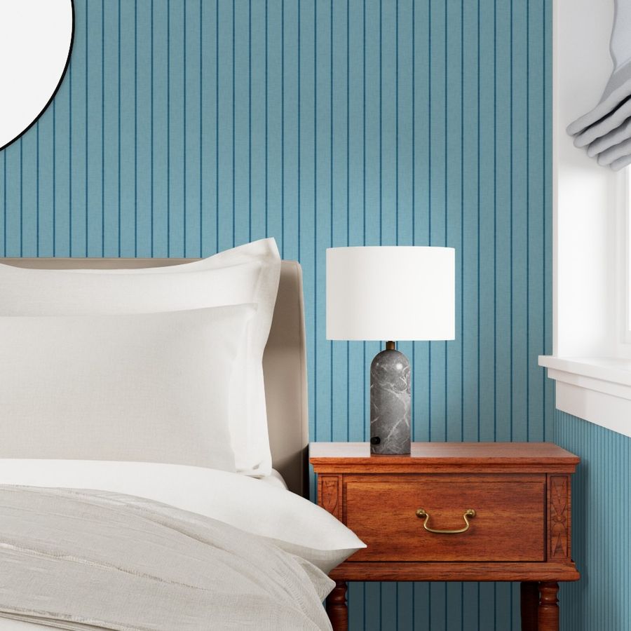 Soft blue Roselk Design wallpaper with fine dark blue vertical stripes behind white bedding, a wooden bedside table and lamp.