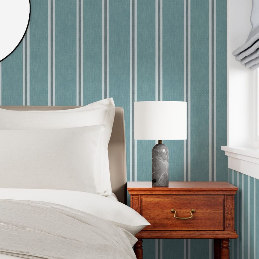 Muted blue Nantucket Linen Textured Stripe wallpaper by Roselk Design behind a bed and wooden bedside table with lamp.